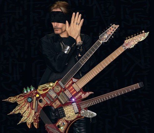 Blindfolded Steve Vai holding the Hydra, a chimeric guitar with two necks, an extra neck for bass strings, and numerous buttons, dials and lights.